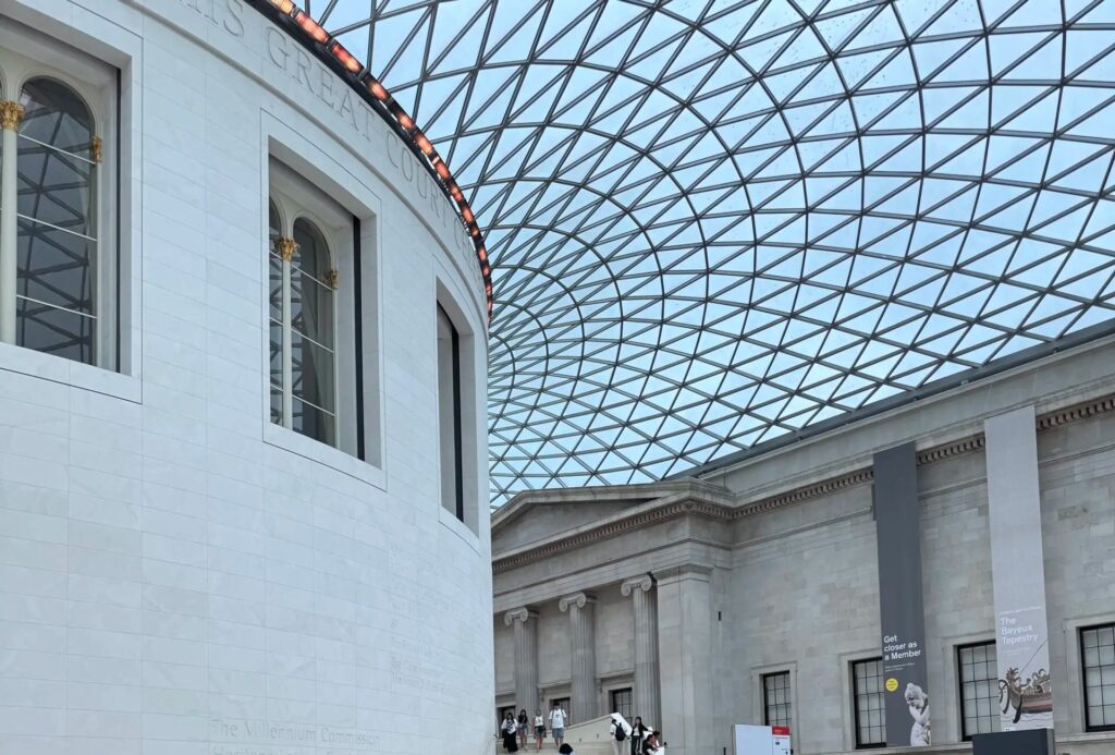 A view of the Queen Elizabeth II Great Court at the British Museum, featuring its iconic glass and steel lattice roof, the circular Reading Room building, and visitors walking toward the museum galleries.