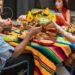Friends passing bowls of food family-style across a colorful dinner table